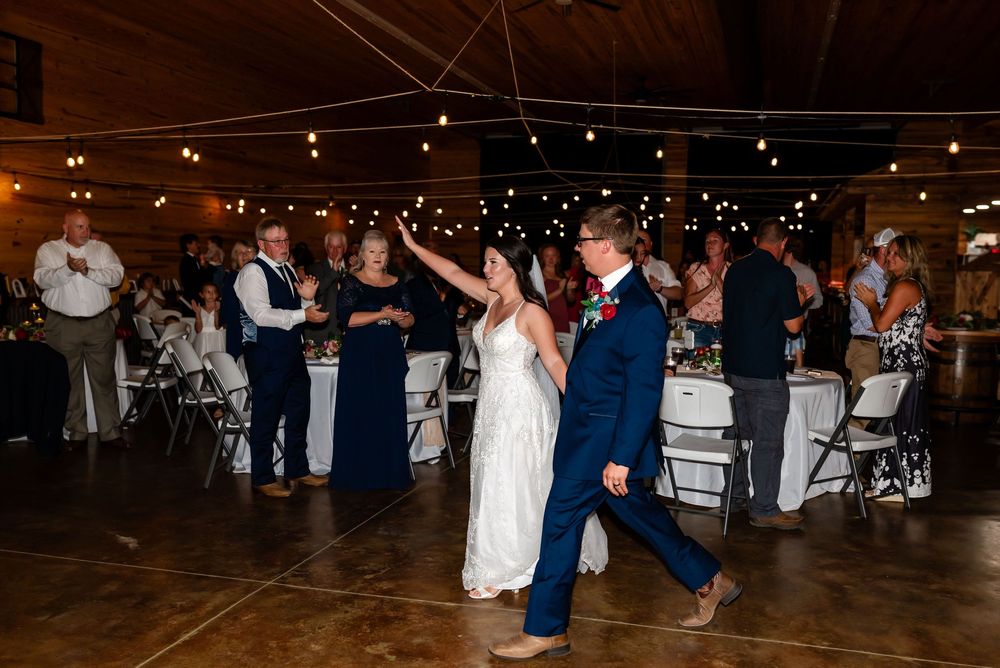 Bride and groom share first dance in rustic barn reception hall with string lights and guests.