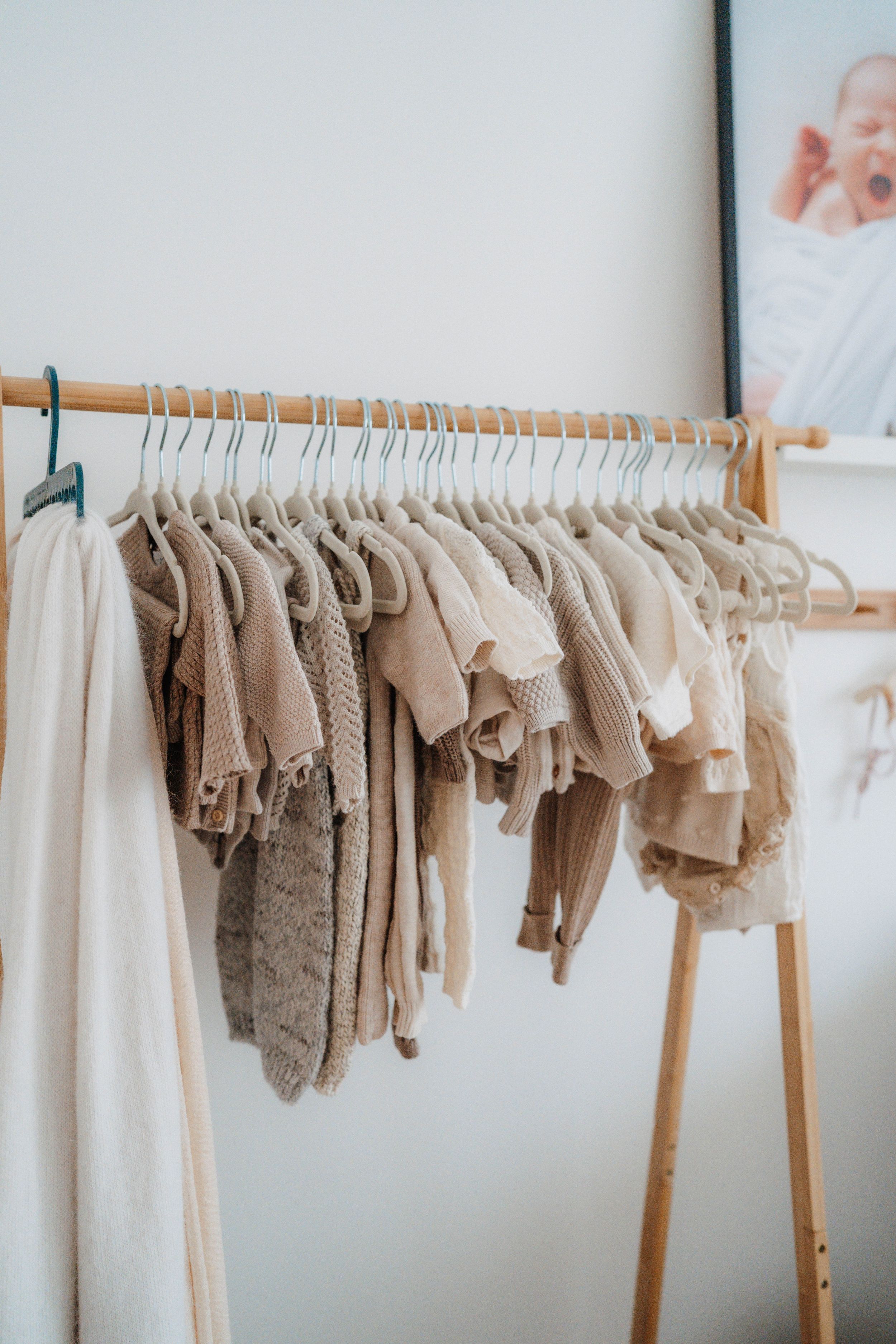 Neutral-toned baby clothes hanging on a wooden clothing rack with white hangers in a minimalist nursery.