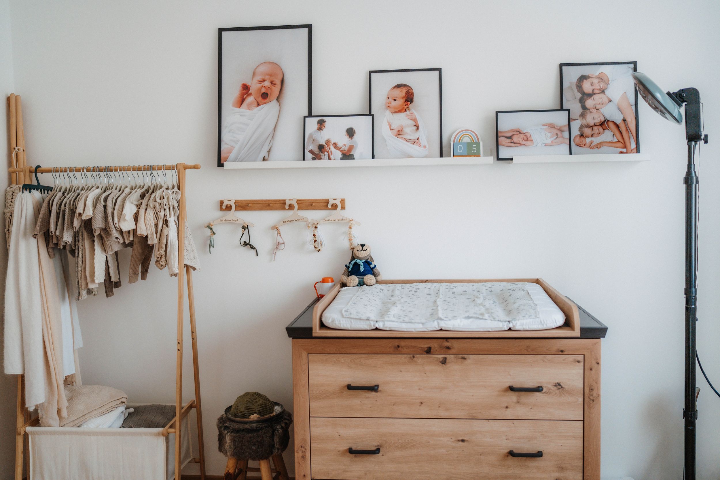 Cozy nursery with wooden dresser changing table, framed baby photos on shelf, and neutral-toned clothing rack.