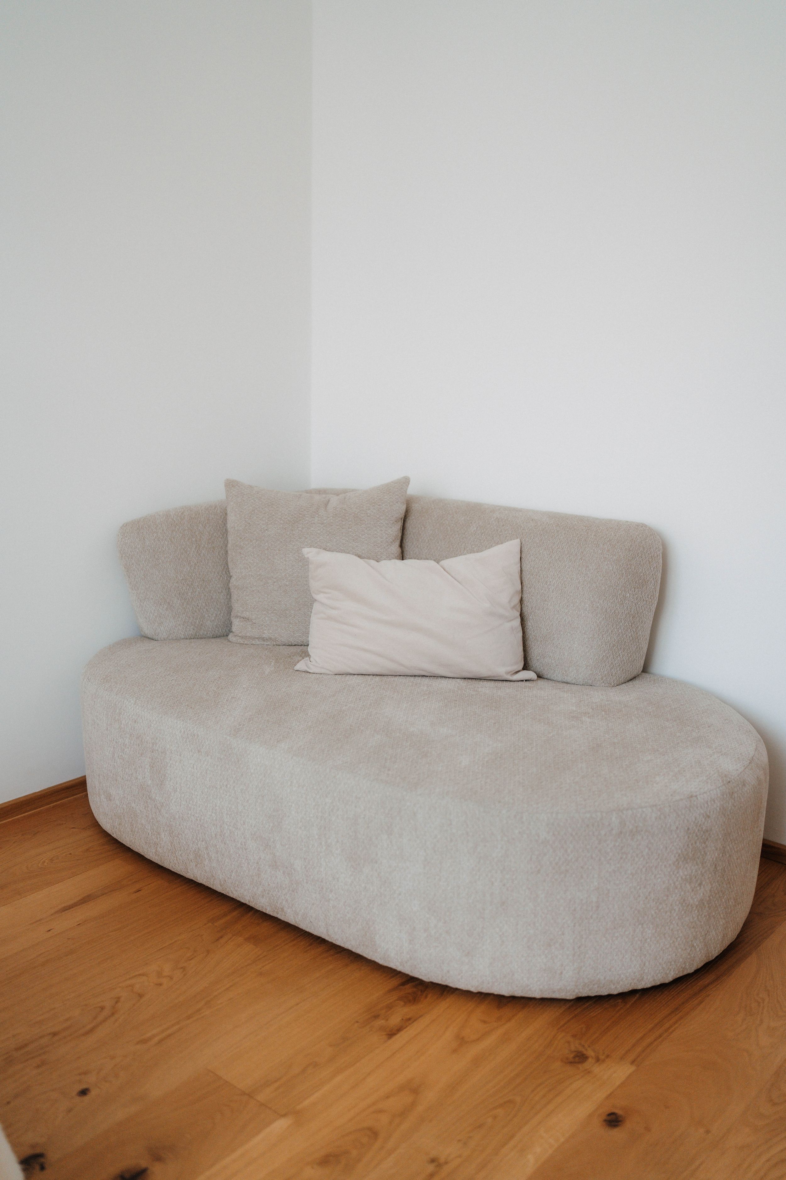 Modern oval beige sofa with cushions on hardwood floor against white walls in a minimalist room.