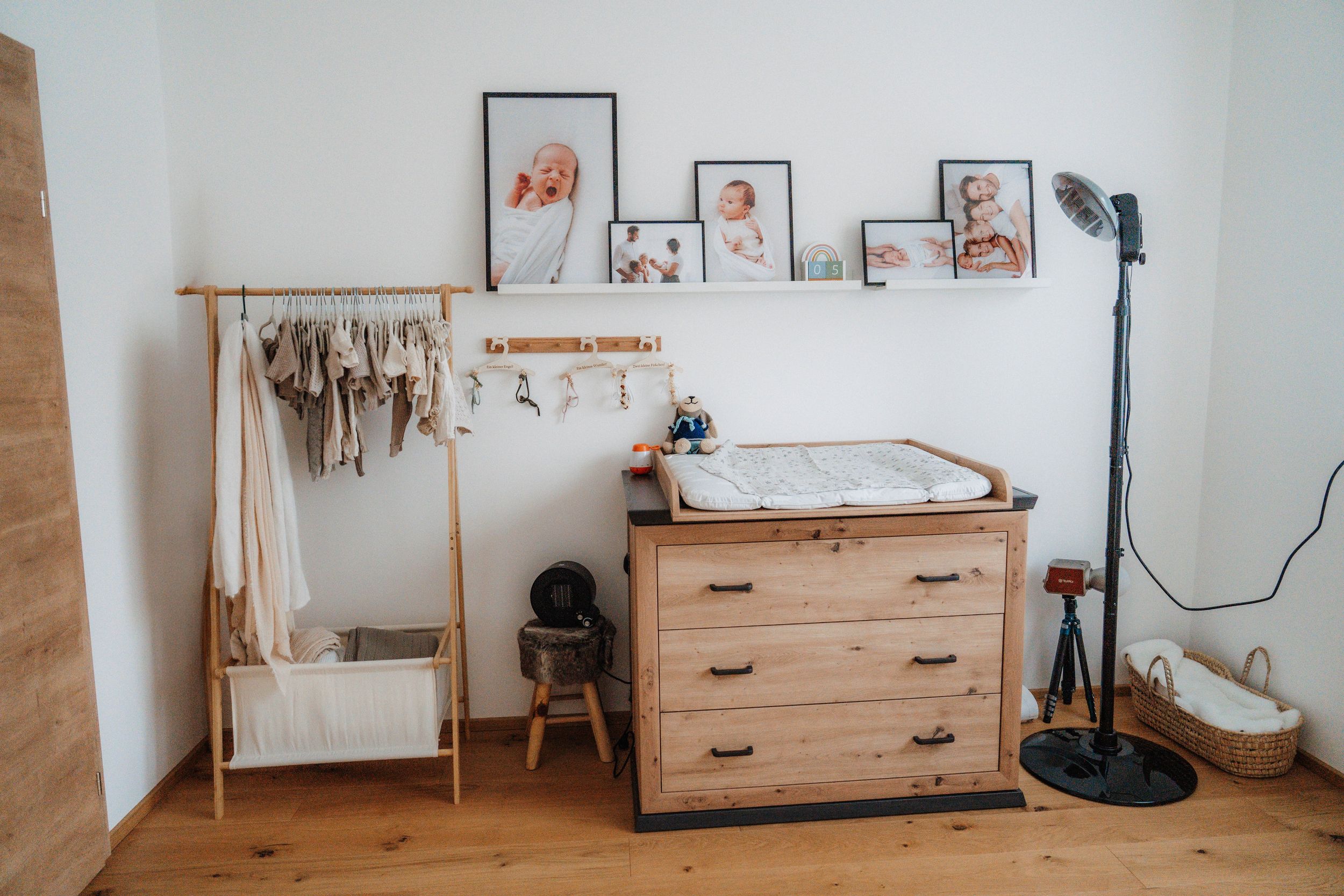 Wooden nursery dresser with changing top, clothing rack, and baby photos on shelf in a bright, modern room.