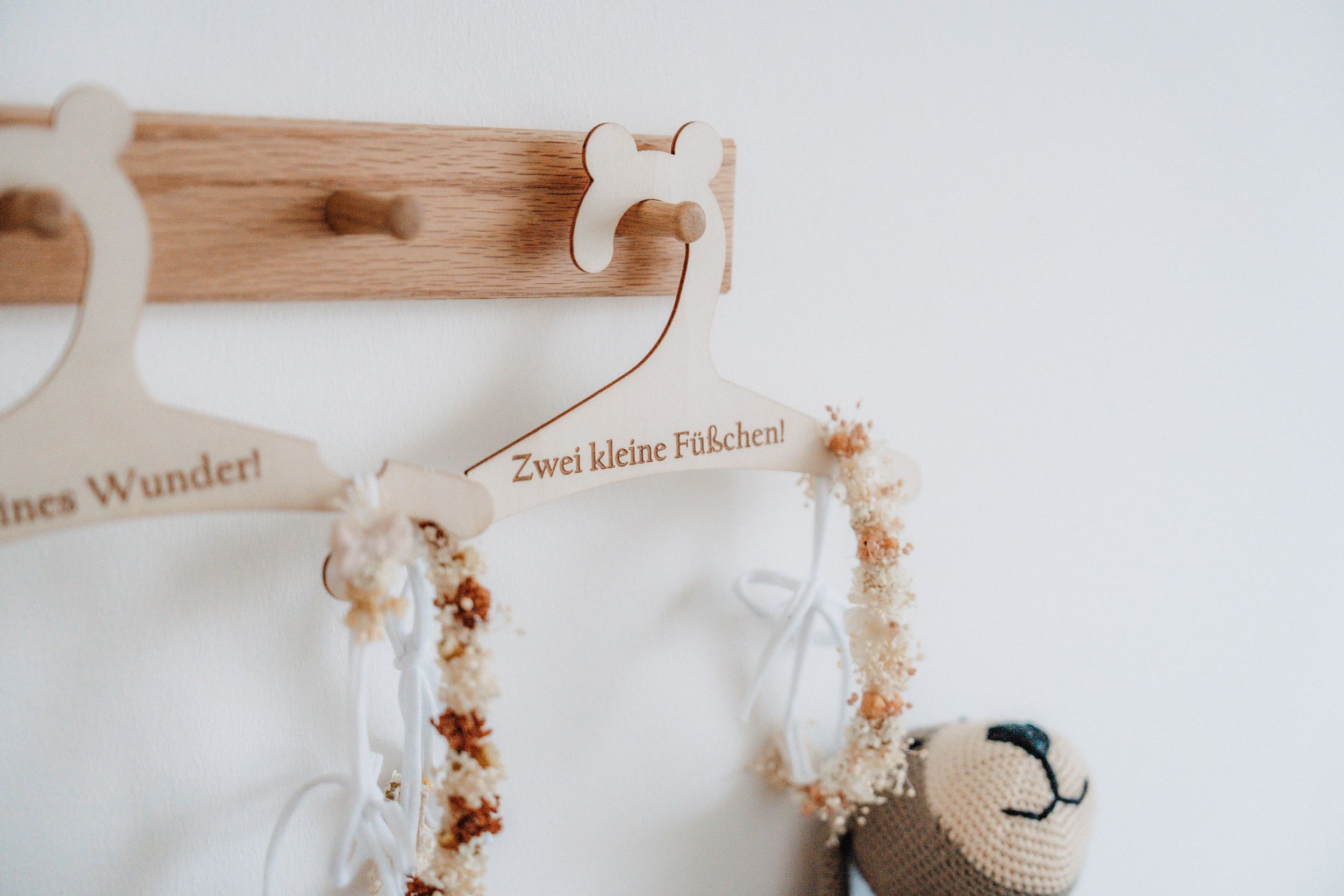 Wooden baby clothes hangers with German text on a wall-mounted coat rack with dried flower garlands.