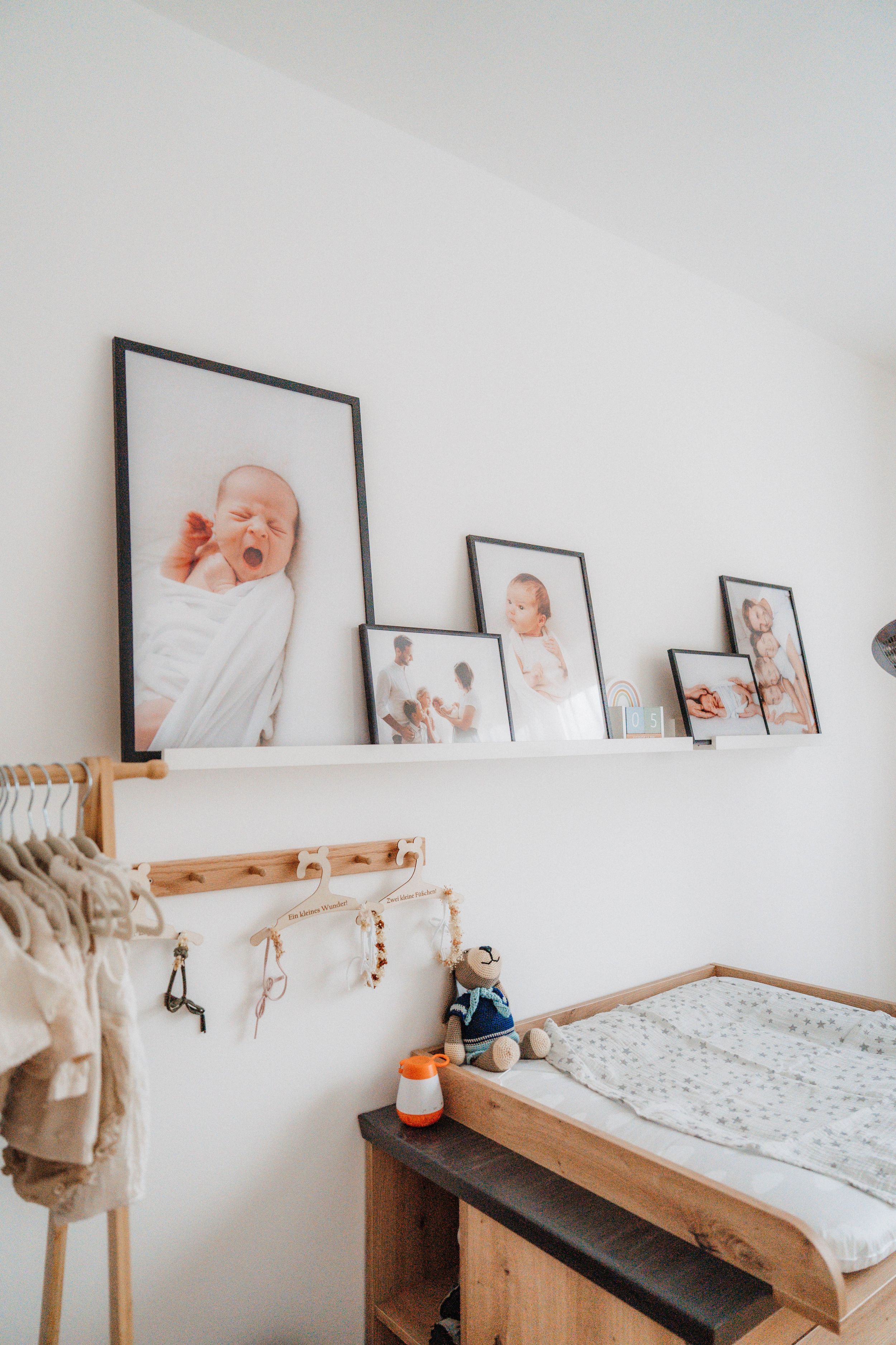 Nursery changing table with wooden dresser, framed newborn photos on white wall shelf, and hanging baby clothes rack.