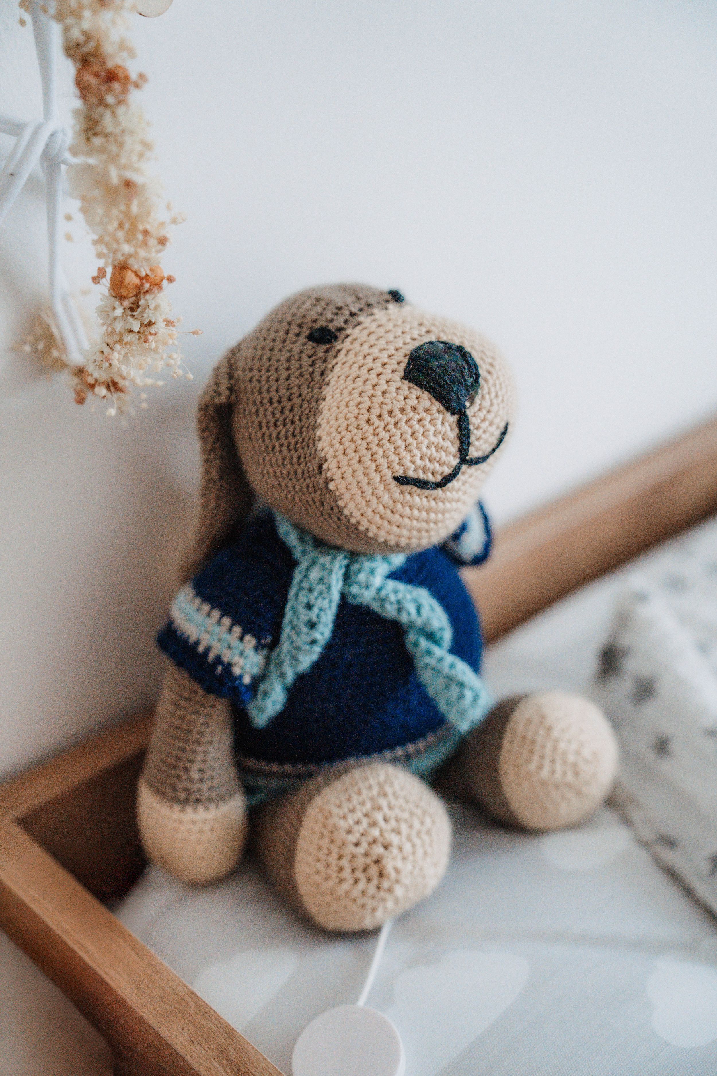 Handmade crochet teddy bear toy in blue sweater sitting on wooden crib in a baby nursery room.