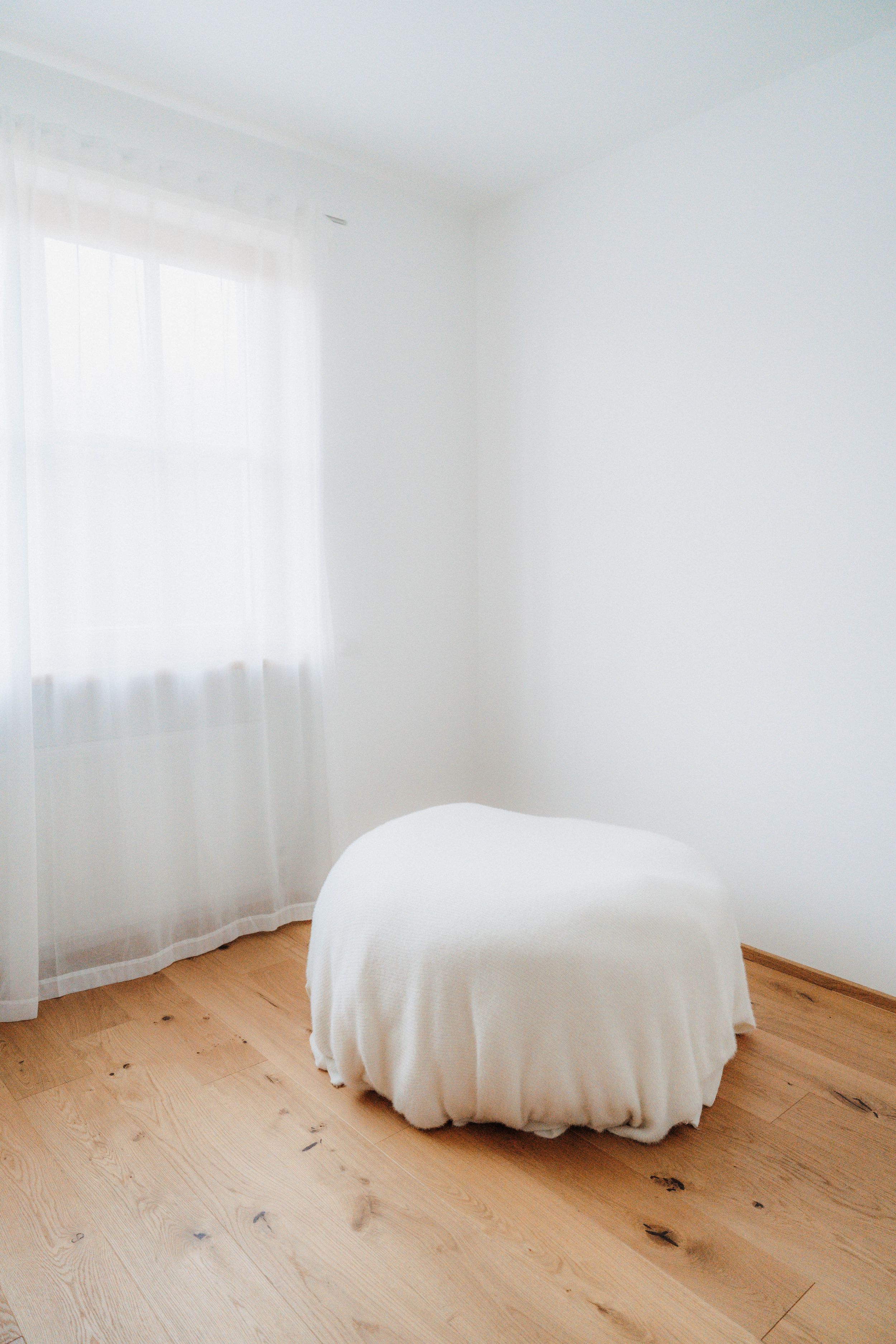 A minimalist white room with sheer curtains, hardwood floors, and a small round table covered with a white cloth.