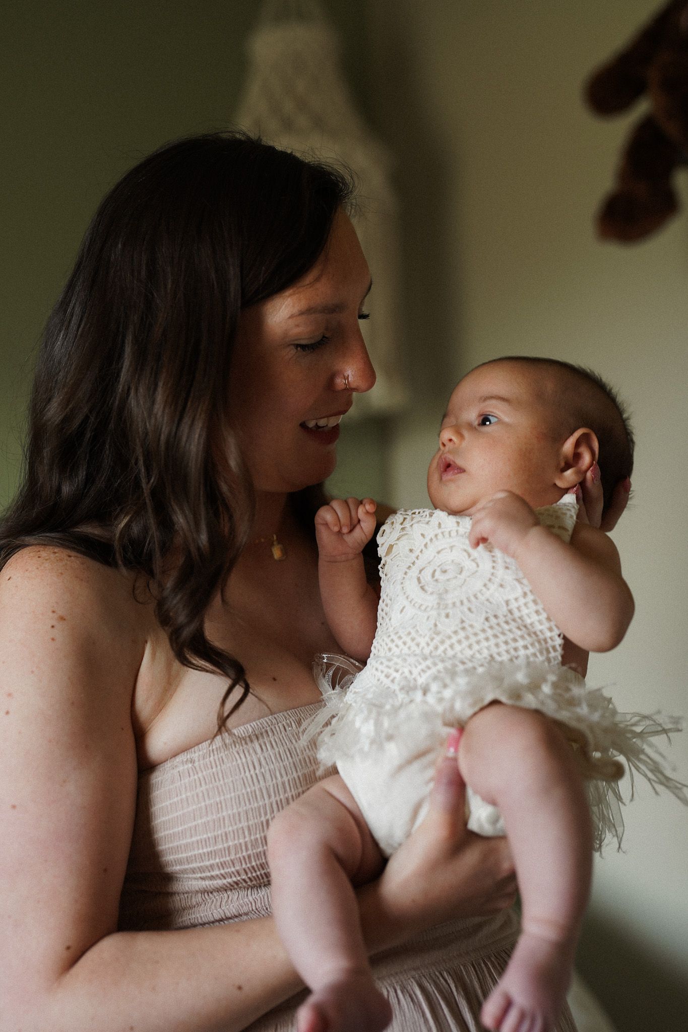 A smiling mother holds her newborn baby dressed in a white crochet outfit, sharing a tender moment together.