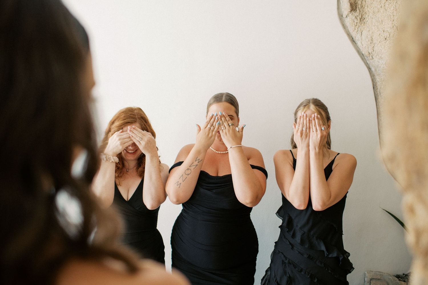 Three bridesmaids in black dresses covering their eyes with hands during a wedding reveal moment.
