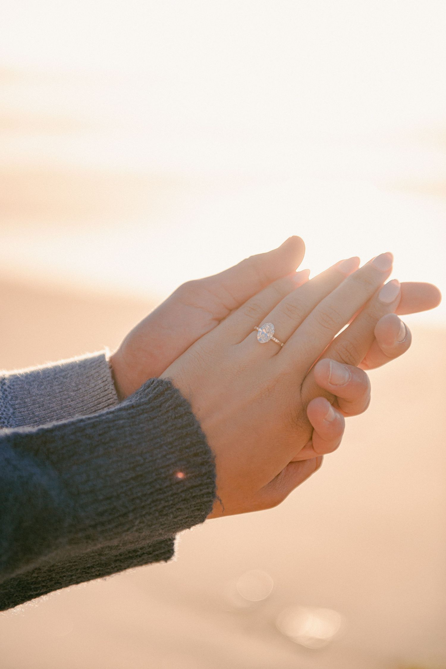 Two hands clasped together on a beach at sunset, showcasing a diamond engagement ring on the ring finger.