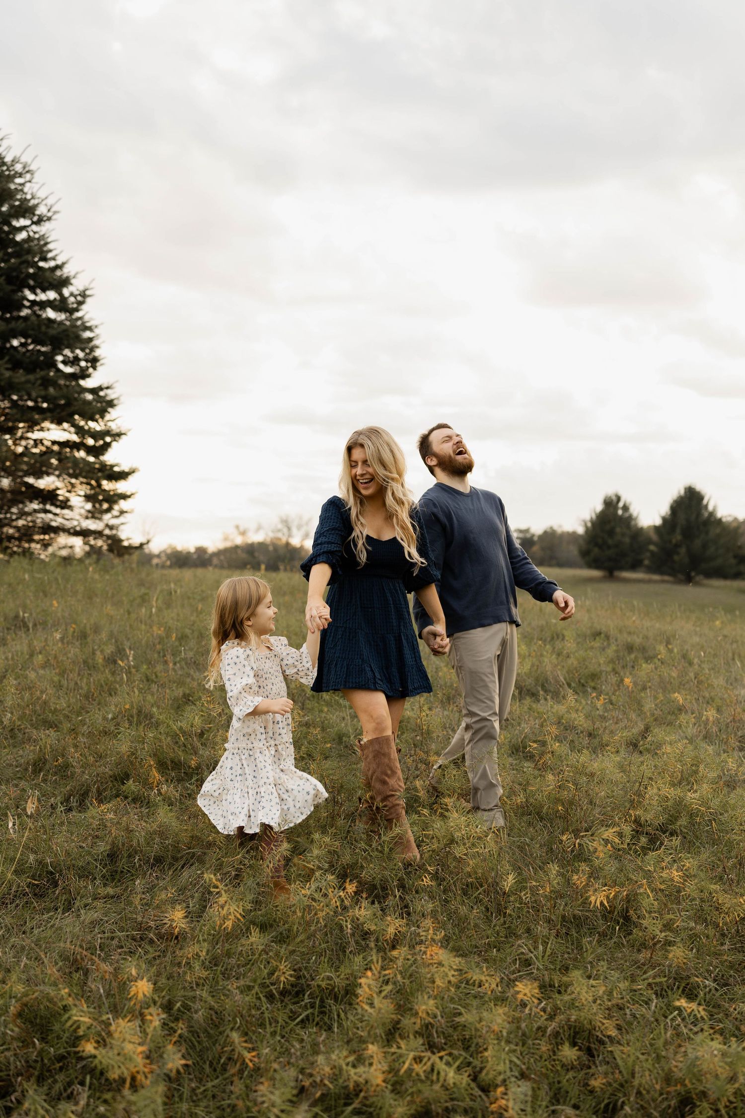 A family of three walks through a green field, with a woman in a navy dress, a bearded man, and a young girl in a floral dress.