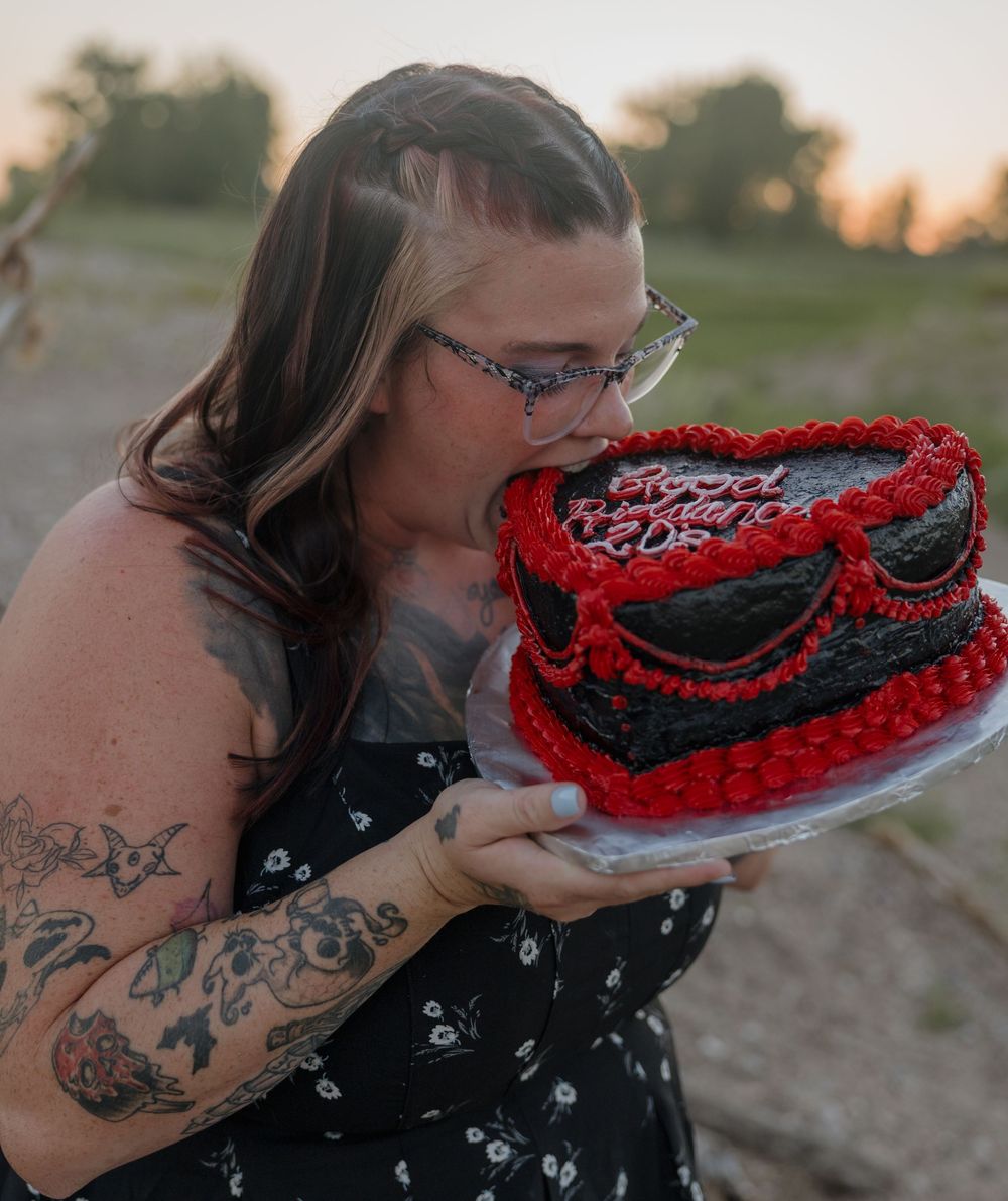 Woman with tattoos kissing a red and black heart-shaped birthday cake outdoors at sunset.