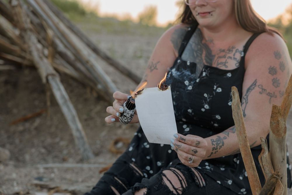 Tattooed woman in black floral dress burning paper outdoors, sitting on chair near driftwood structure.