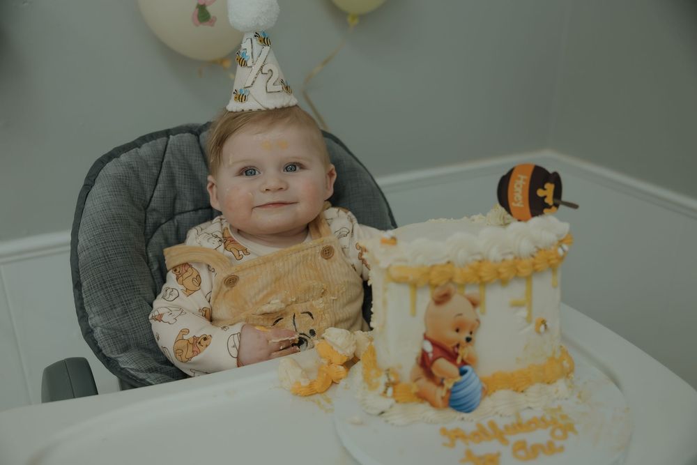 Baby in high chair wearing party hat celebrates first birthday with Winnie the Pooh themed smash cake.