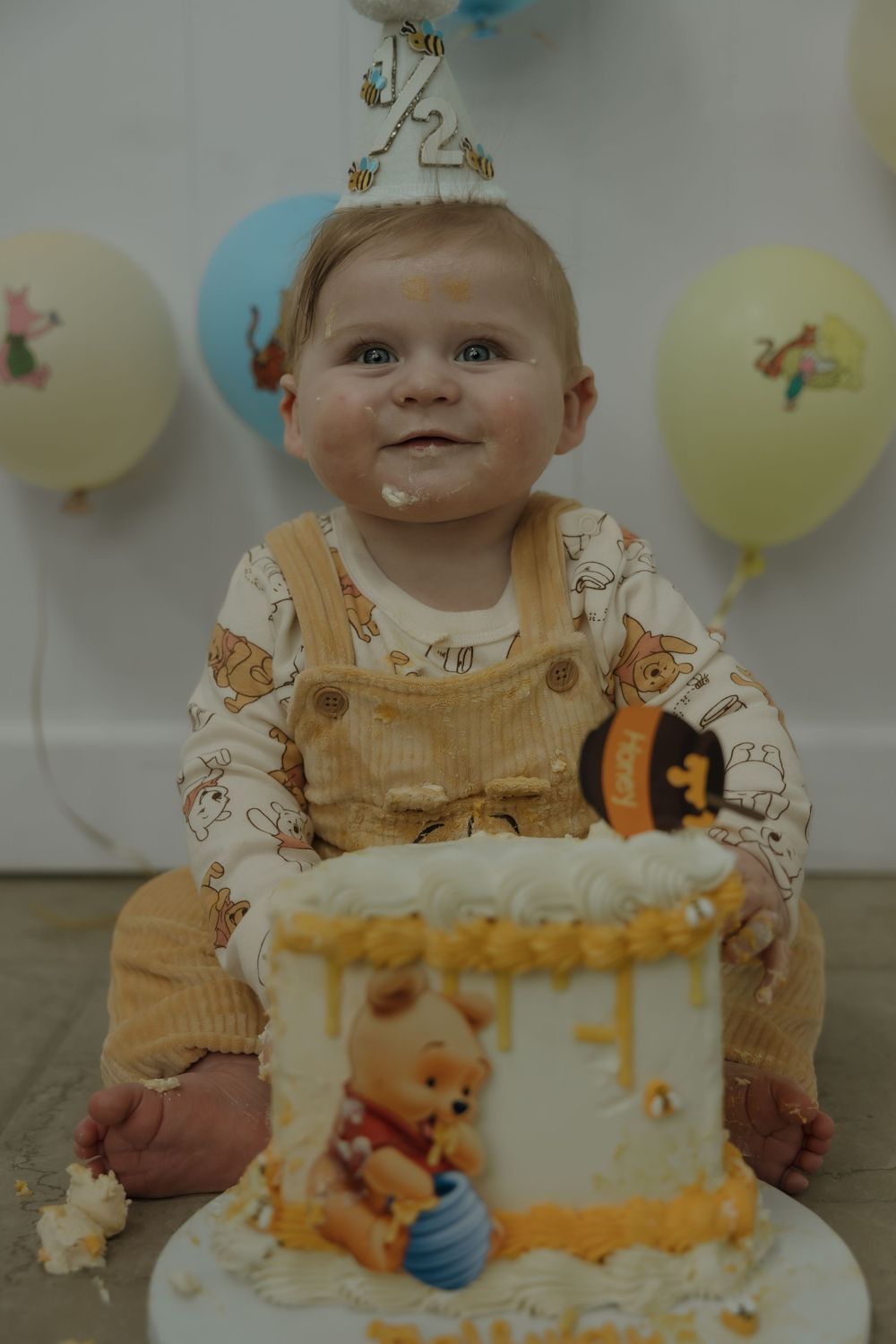 Baby smiling at first birthday cake smash with Winnie the Pooh themed decorations, balloons, and honey drip cake.