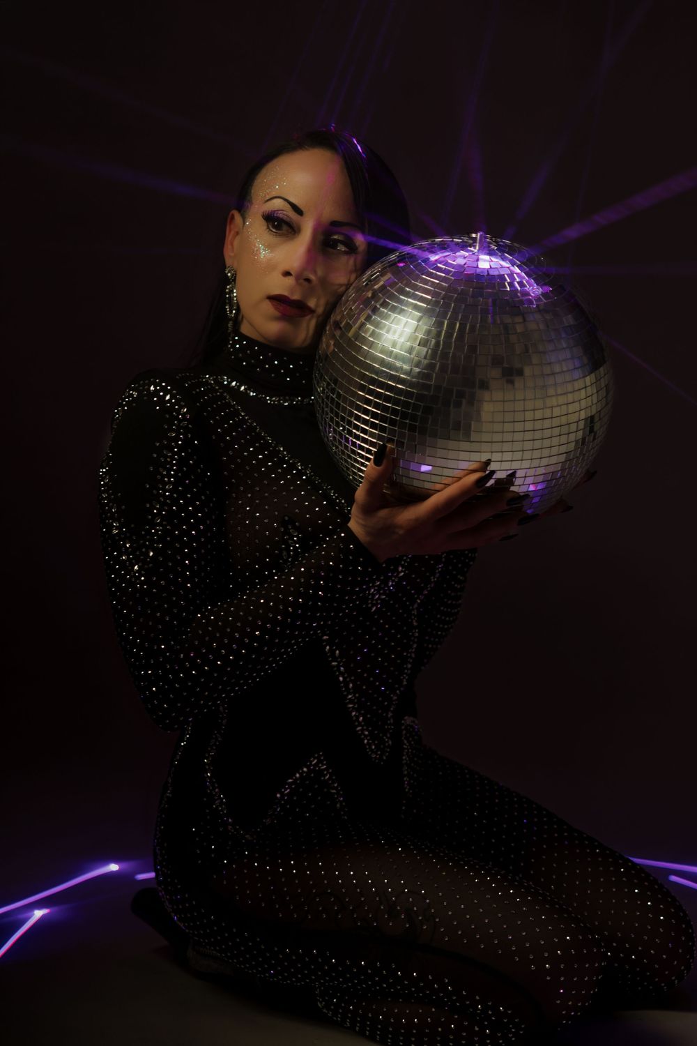Woman in black rhinestone bodysuit holding a shiny disco ball with purple laser lights in dark studio.