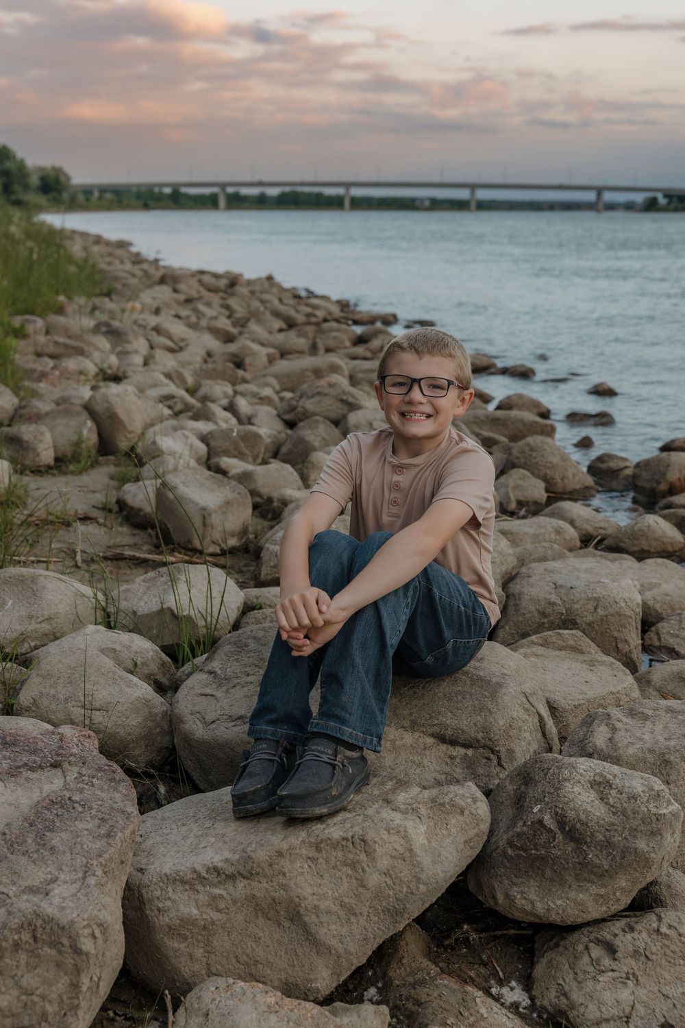 Smiling boy with glasses sitting on rocky riverbank at sunset wearing jeans and a pink shirt.