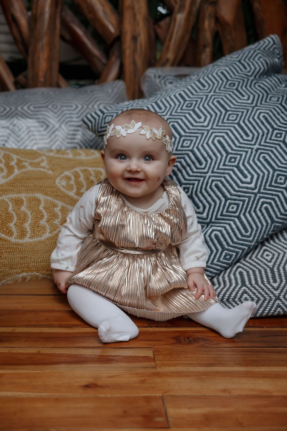 Smiling baby girl in gold dress and floral headband sitting on wooden floor surrounded by decorative pillows. Bismarck, ND photographer