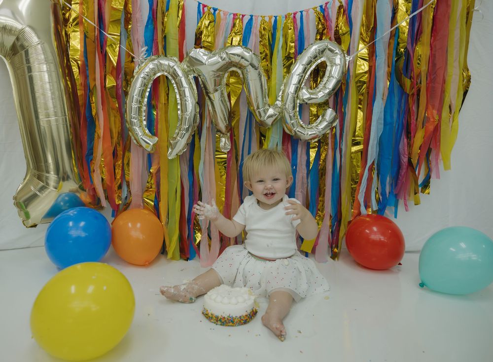 Baby's first birthday cake smash with colorful balloons and silver 'one' balloon letters on gold fringe backdrop. Bismarck, ND photographer