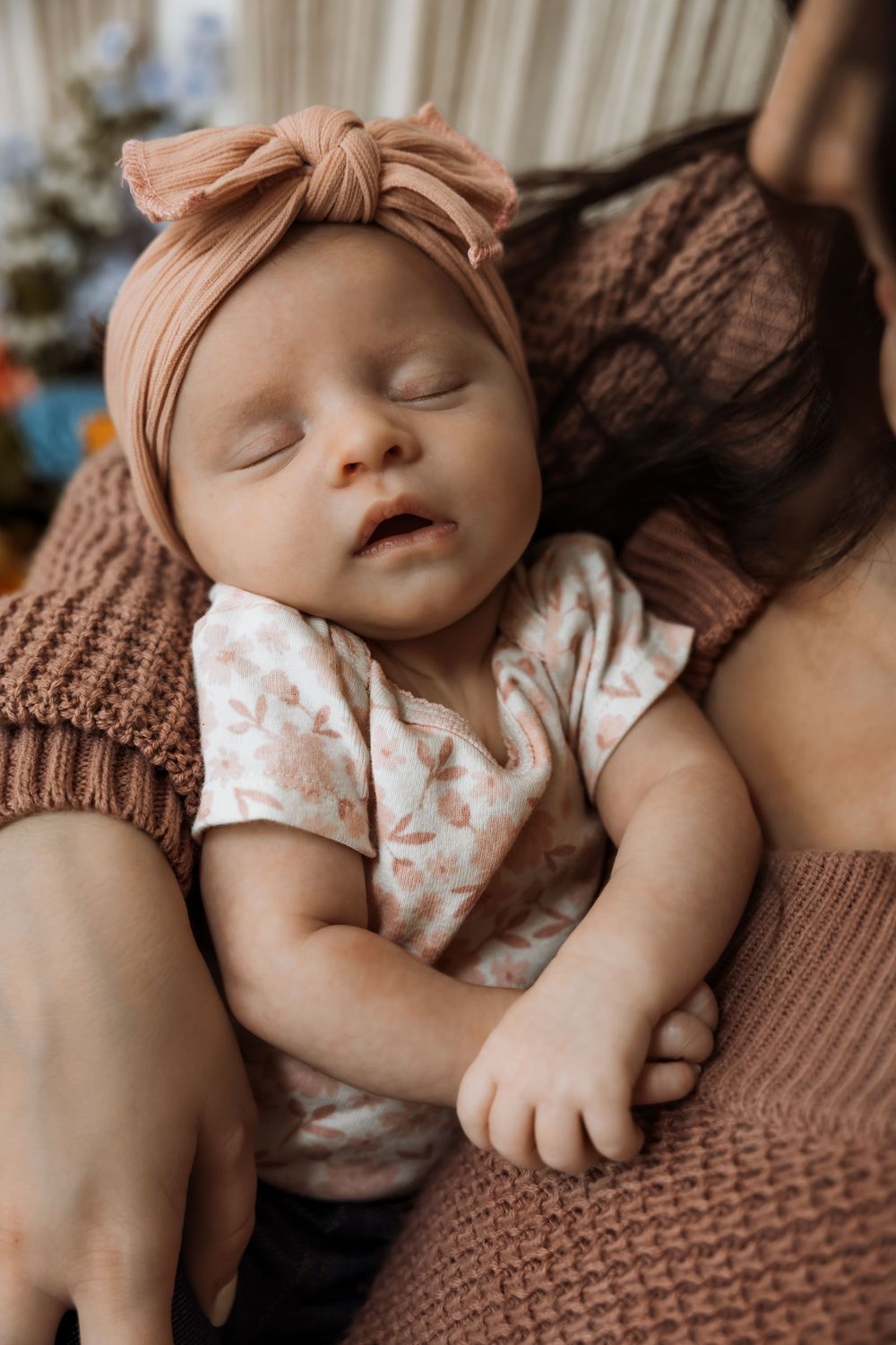 Sleeping newborn baby in floral onesie and pink bow headband resting peacefully in mother's arms. Bismarck, ND photographer