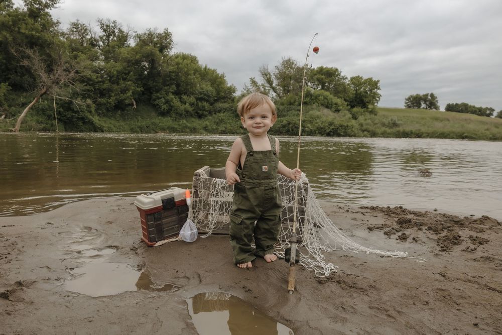 Toddler boy in overalls stands barefoot in muddy river bank holding fishing net with tackle box nearby. Bismarck, ND photographer
