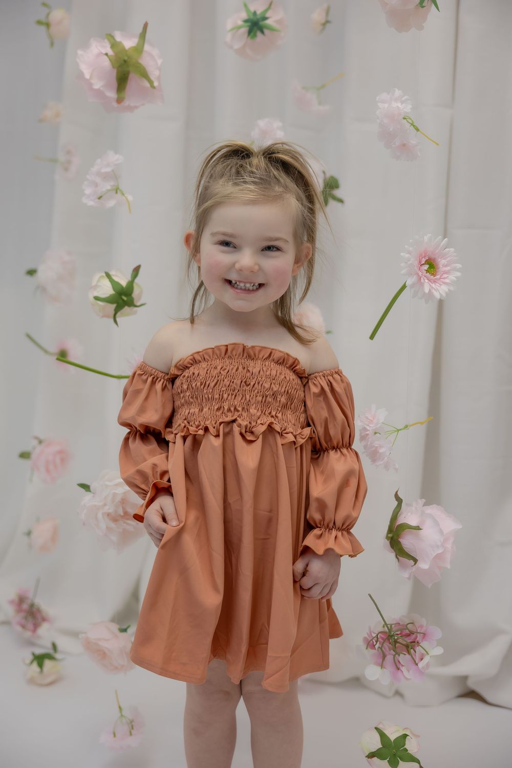 Smiling toddler girl in rust off-shoulder smocked dress poses before floral backdrop with pink blooms. Bismarck, ND photographer