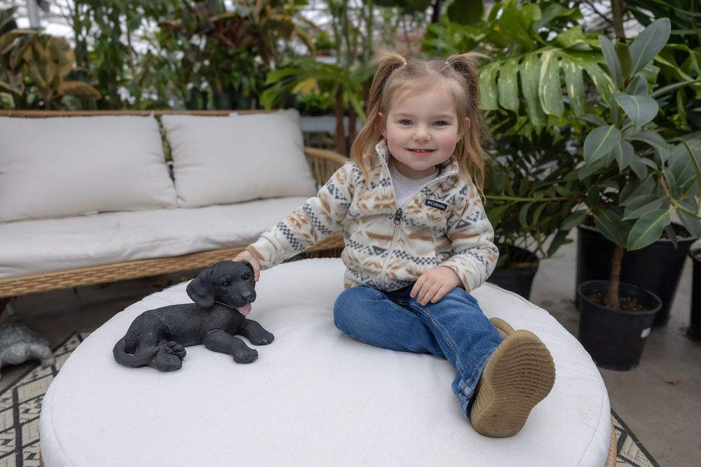 Young girl sitting on a white ottoman with a black puppy figurine, surrounded by lush green plants indoors. Bismarck, ND photographer