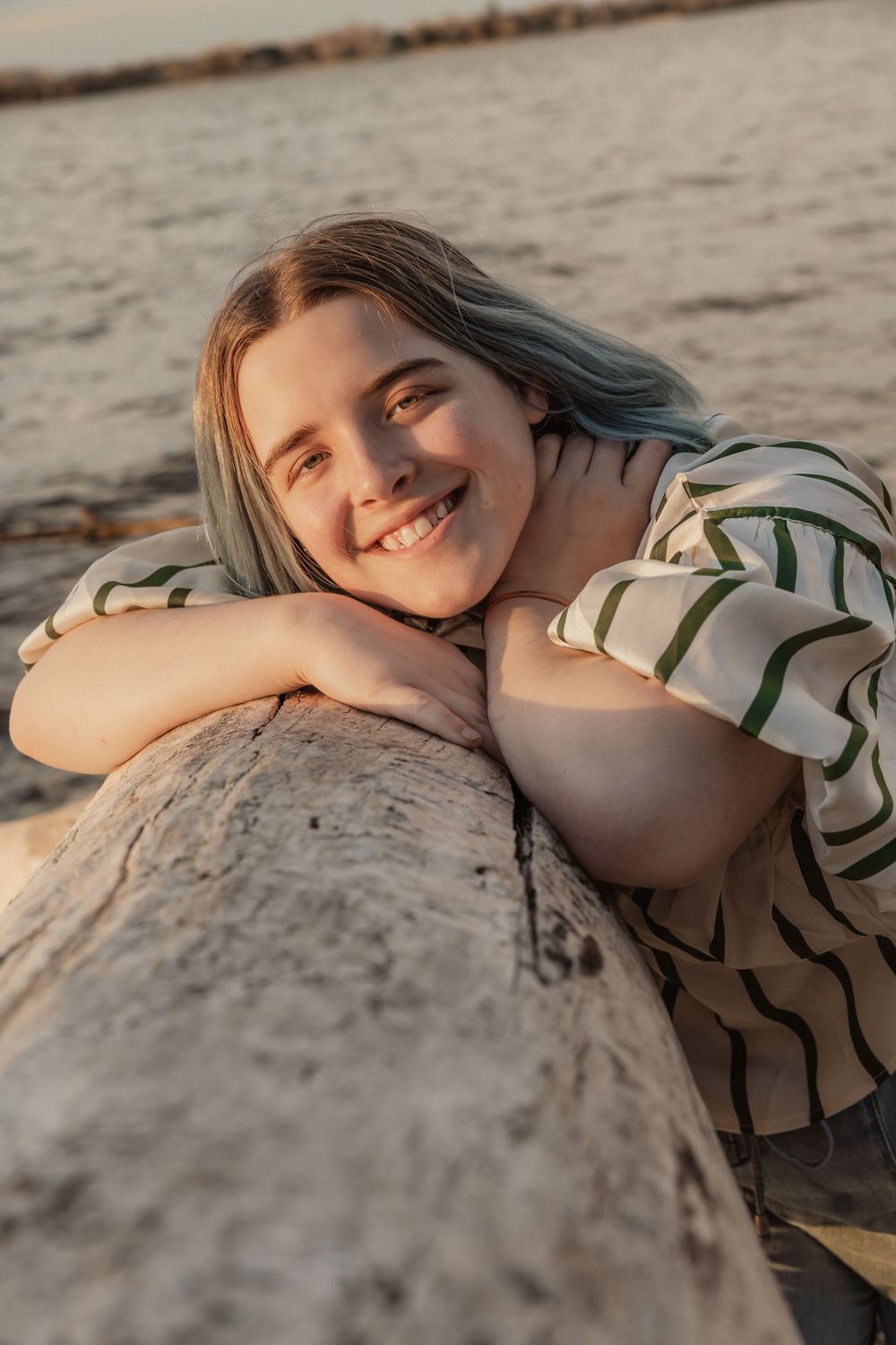 Smiling young woman with blonde hair leaning on a driftwood log at a beach during golden hour sunset. Bismarck, ND photographer