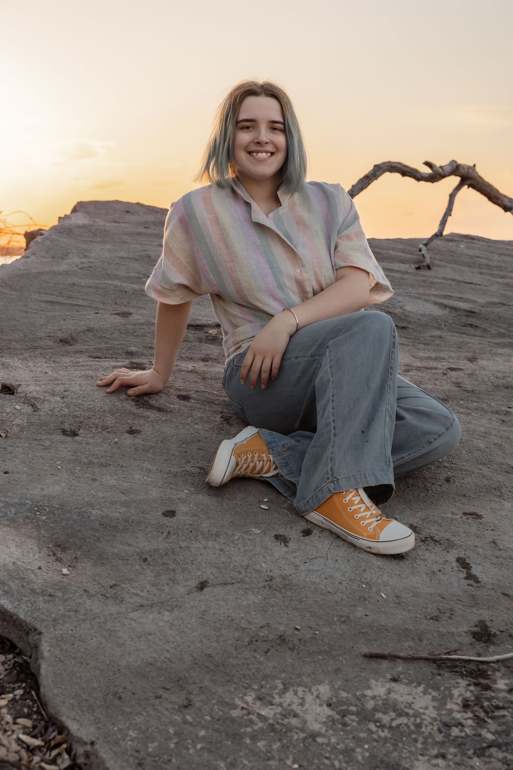 Young woman with blue hair sits on rock at sunset, wearing striped shirt, jeans, and orange Converse sneakers. Bismarck, ND photographer