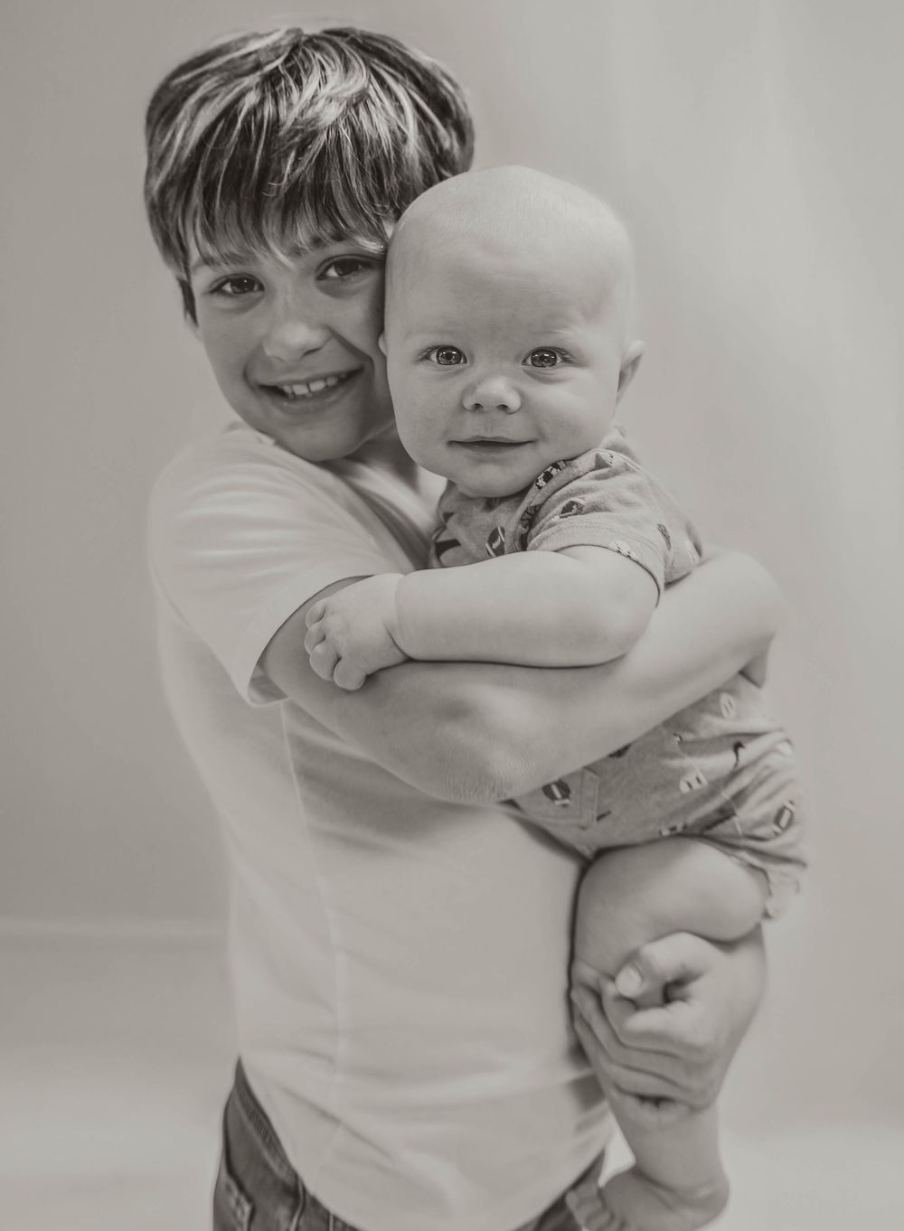 Black and white photo of a smiling boy hugging a cute baby, both looking at the camera in a studio setting. Garrison, ND Photographer