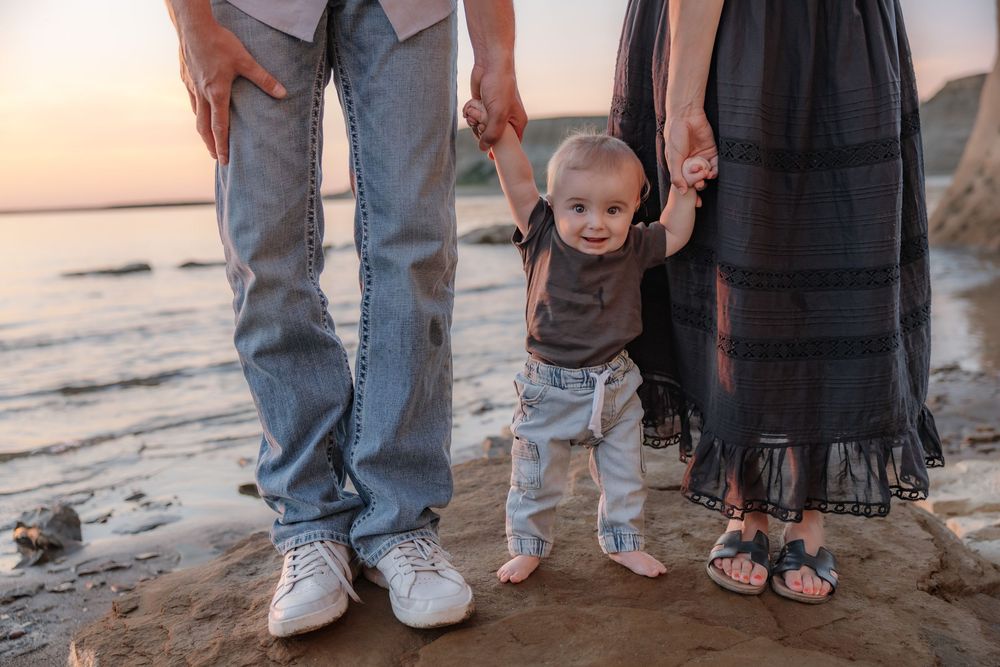 A baby takes first steps on a sandy beach at sunset, held by parents hands on either side. Garrison, ND Photographer