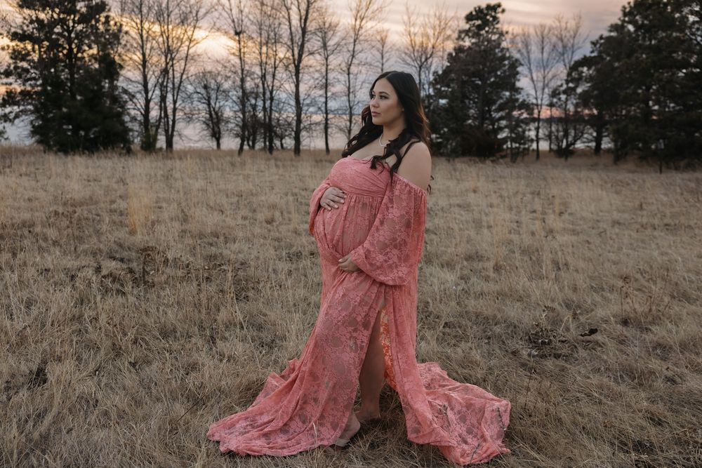 Pregnant woman in pink lace maternity gown standing in a field at sunset with bare trees in the background. Garrison, ND Photographer