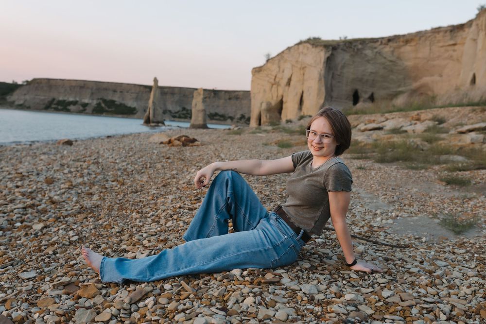 A smiling woman sits relaxed on a rocky beach wearing jeans and a gray tee, with ancient stone ruins and calm water behind her. Garrison, ND