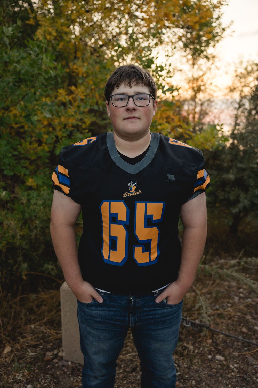 Teen boy in football jersey number 65 standing outdoors among autumn trees at sunset. Garrison, ND Photographer