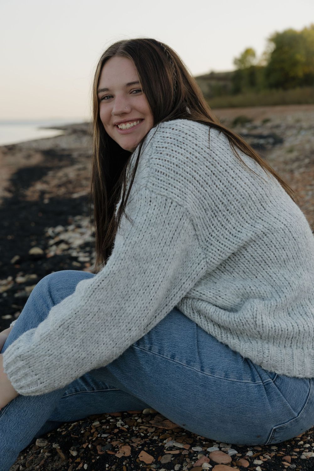 Smiling young woman with long brown hair sitting on a rocky beach wearing a gray knit sweater and blue jeans. Garrison, ND Photographer