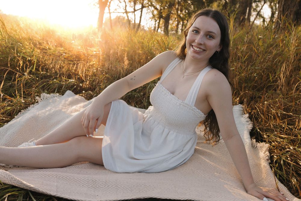 Young woman in white dress sitting on blanket in sunlit field smiling at golden hour. Garrison, ND Photographer