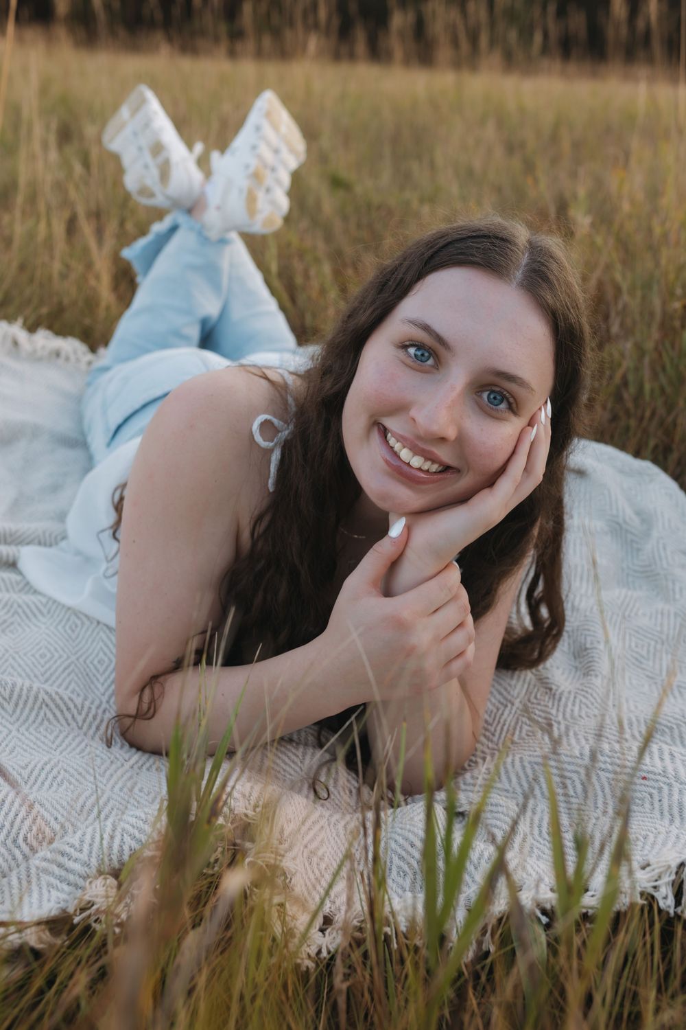 Young woman in light blue dress lying on blanket in golden grass field, smiling with hand on cheek. Garrison, ND Photographer