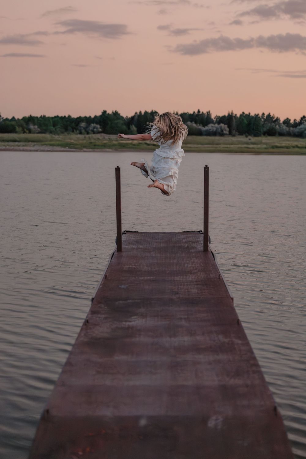 Woman in white dress jumping off a rustic wooden dock over a calm lake at sunset with trees in the background. Travel Photographer