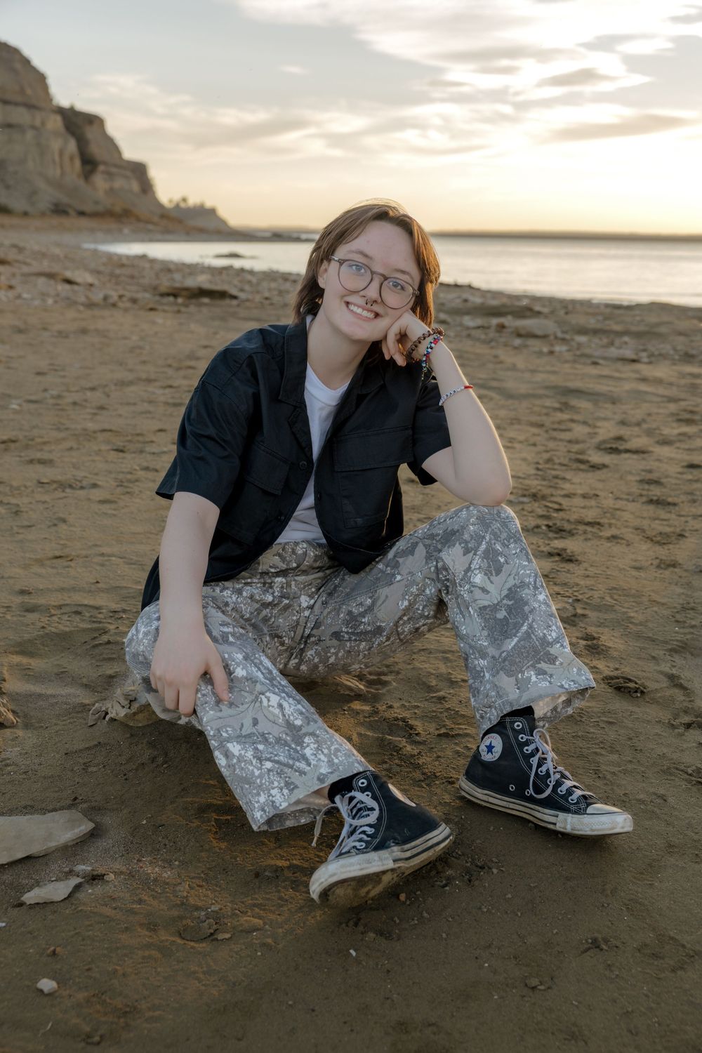 Teen girl in glasses sitting on sandy beach at sunset, wearing black jacket, metallic pants, and Converse sneakers. Garrison, ND Photograph