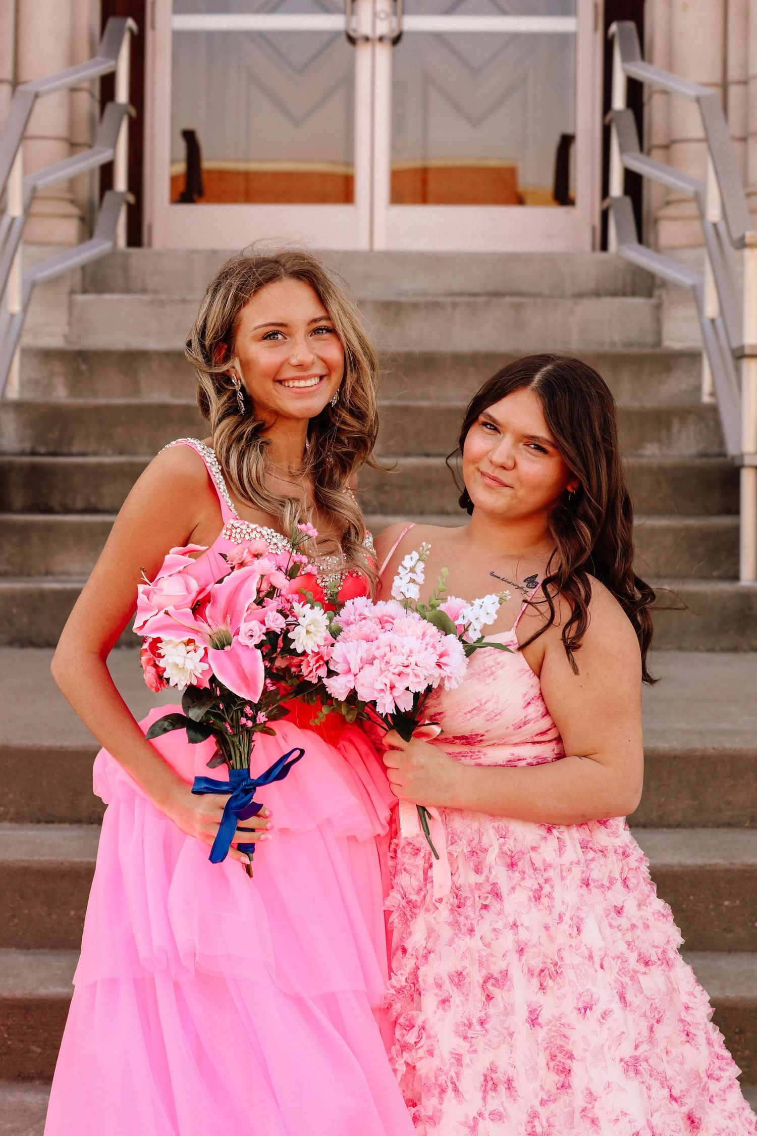Two young women in pink formal dresses holding flower bouquets on steps of a building.