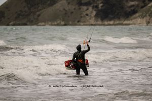 Kitesurfer in wetsuit stands in choppy ocean waves holding kite lines, with rocky hills in the background.