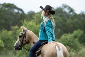 Blonde woman in teal jacket and black cowboy hat riding a palomino horse outdoors with green trees behind.