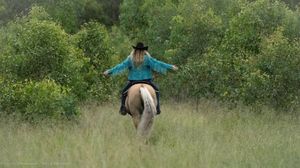 Woman in turquoise shirt riding a palomino horse through tall green grass with lush trees in the background.