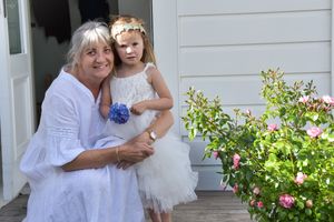 Woman and young girl in white dresses posing together outdoors near pink roses.