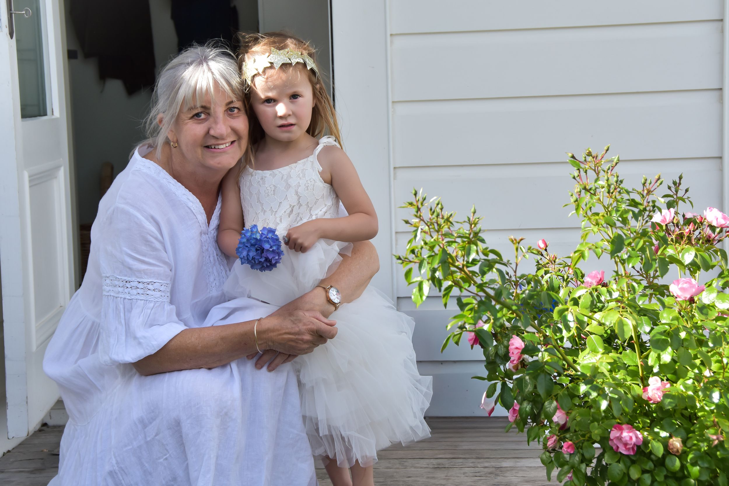 Woman and young girl in white dresses posing together outdoors near pink roses.