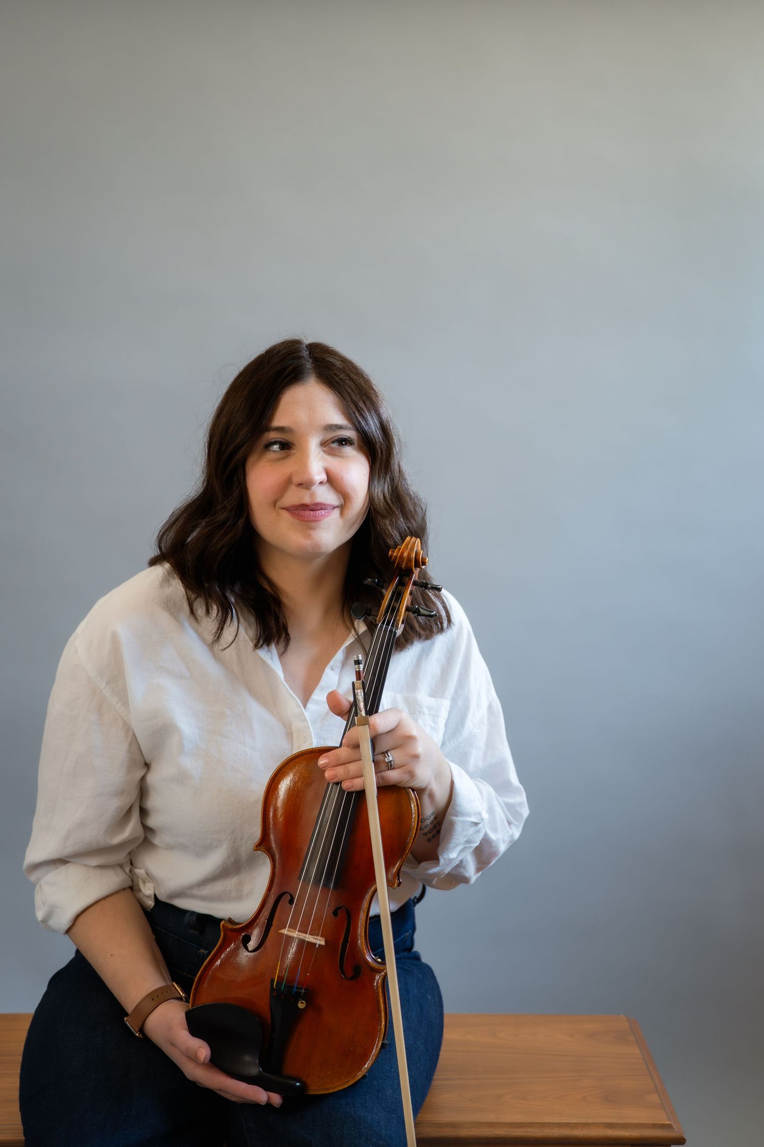 Woman in white blouse holding a violin and bow, smiling thoughtfully while seated against a gray background.