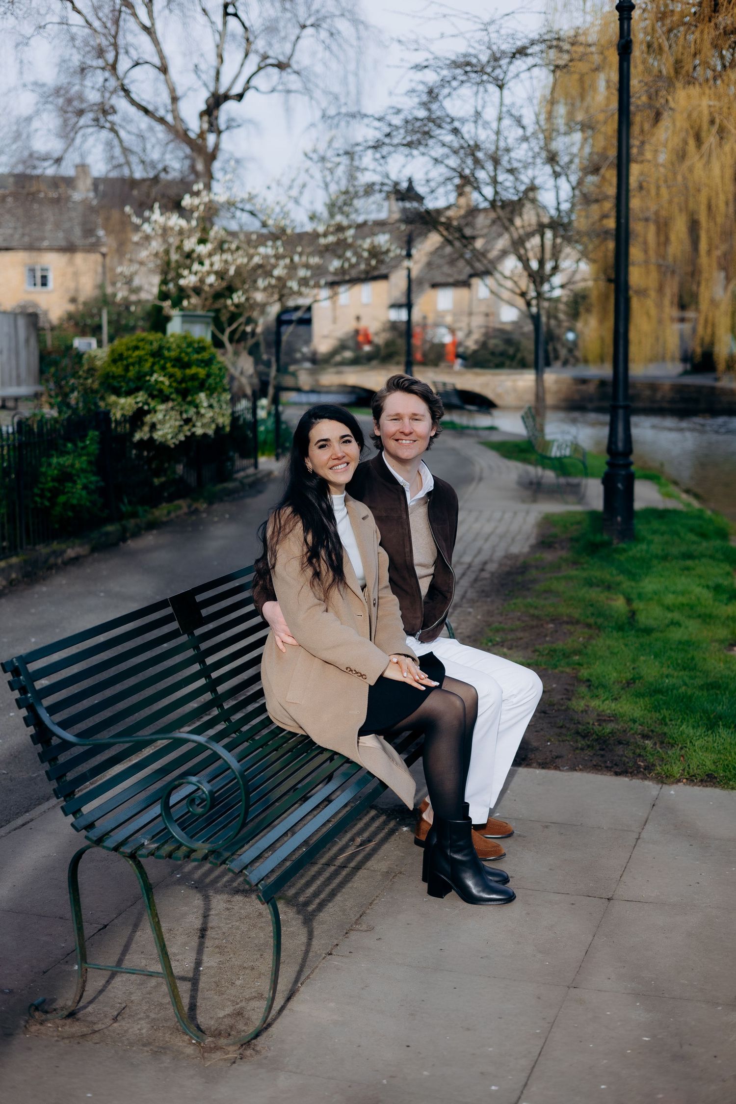 A couple sits smiling on a green bench in bourton on the water.