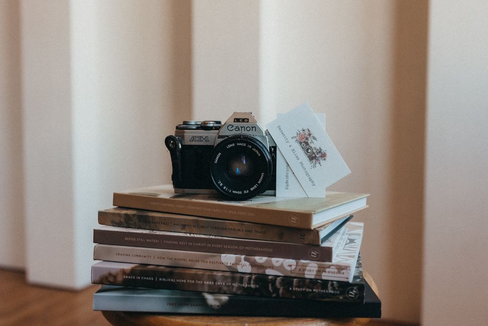 A vintage Canon film camera resting on a stack of books and magazines on a small table indoors.