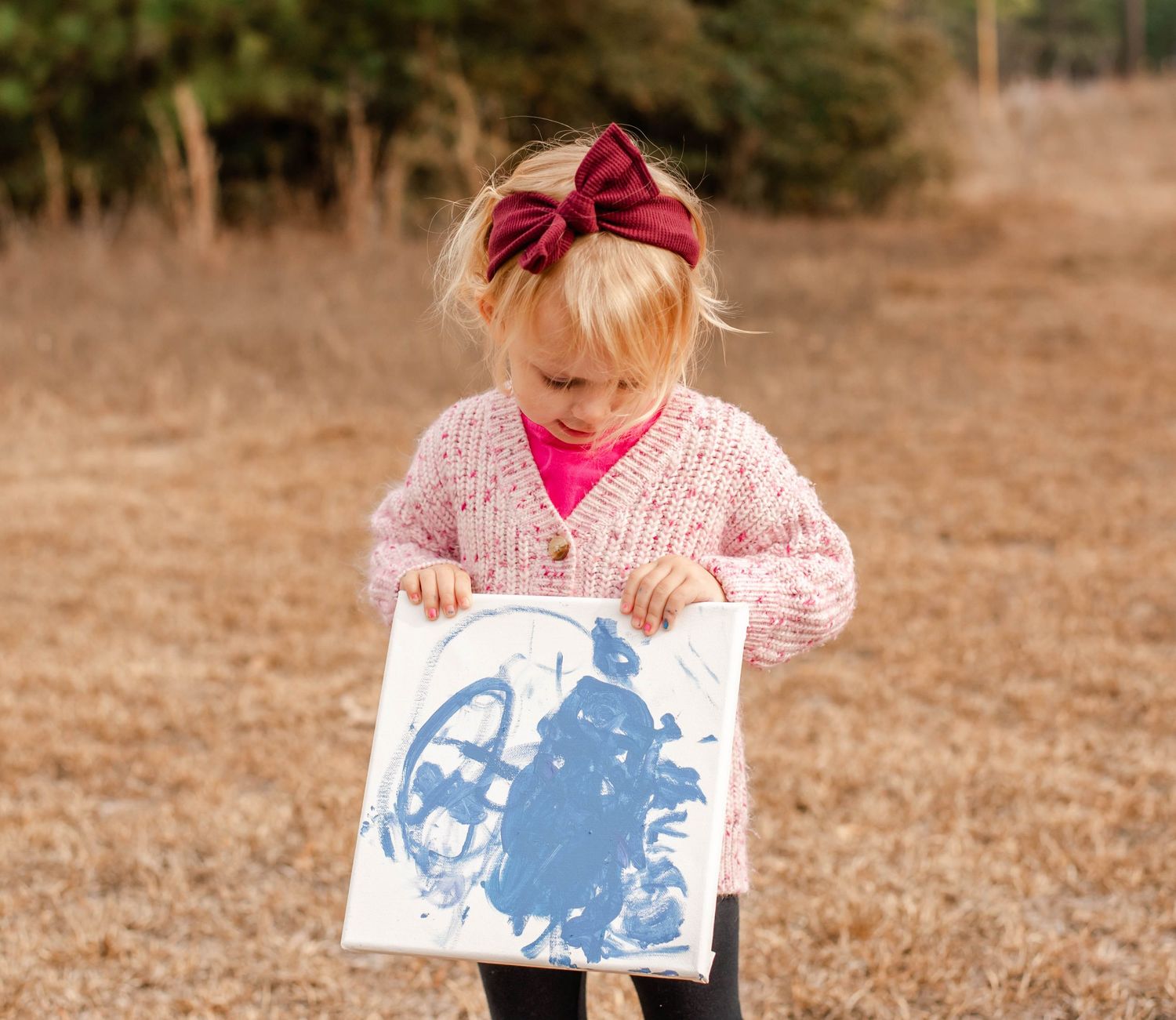 A young girl with a red bow headband holds a blue painting outdoors in a field.