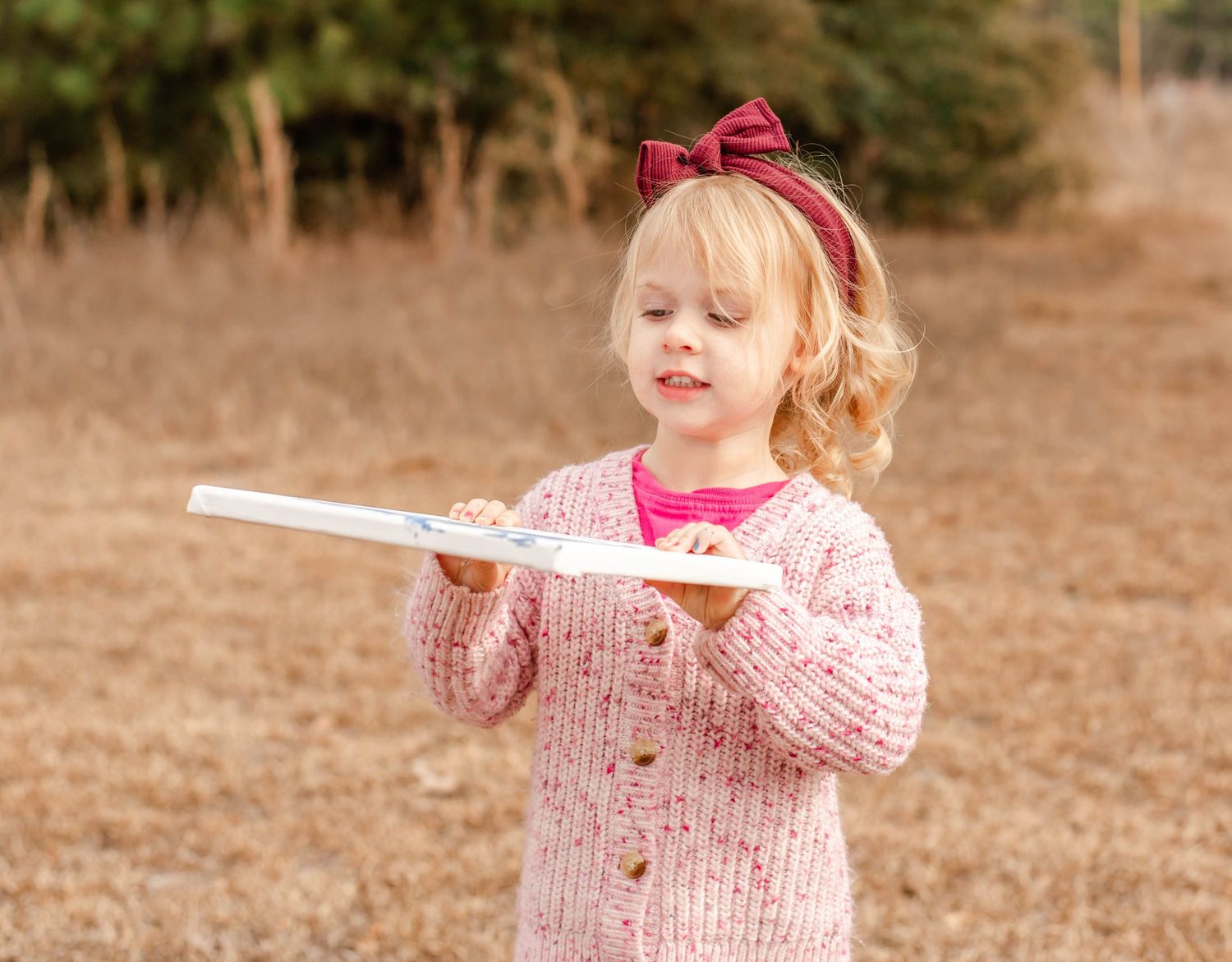 Young girl in pink sweater and red bow headband holds a book outdoors in a natural setting.