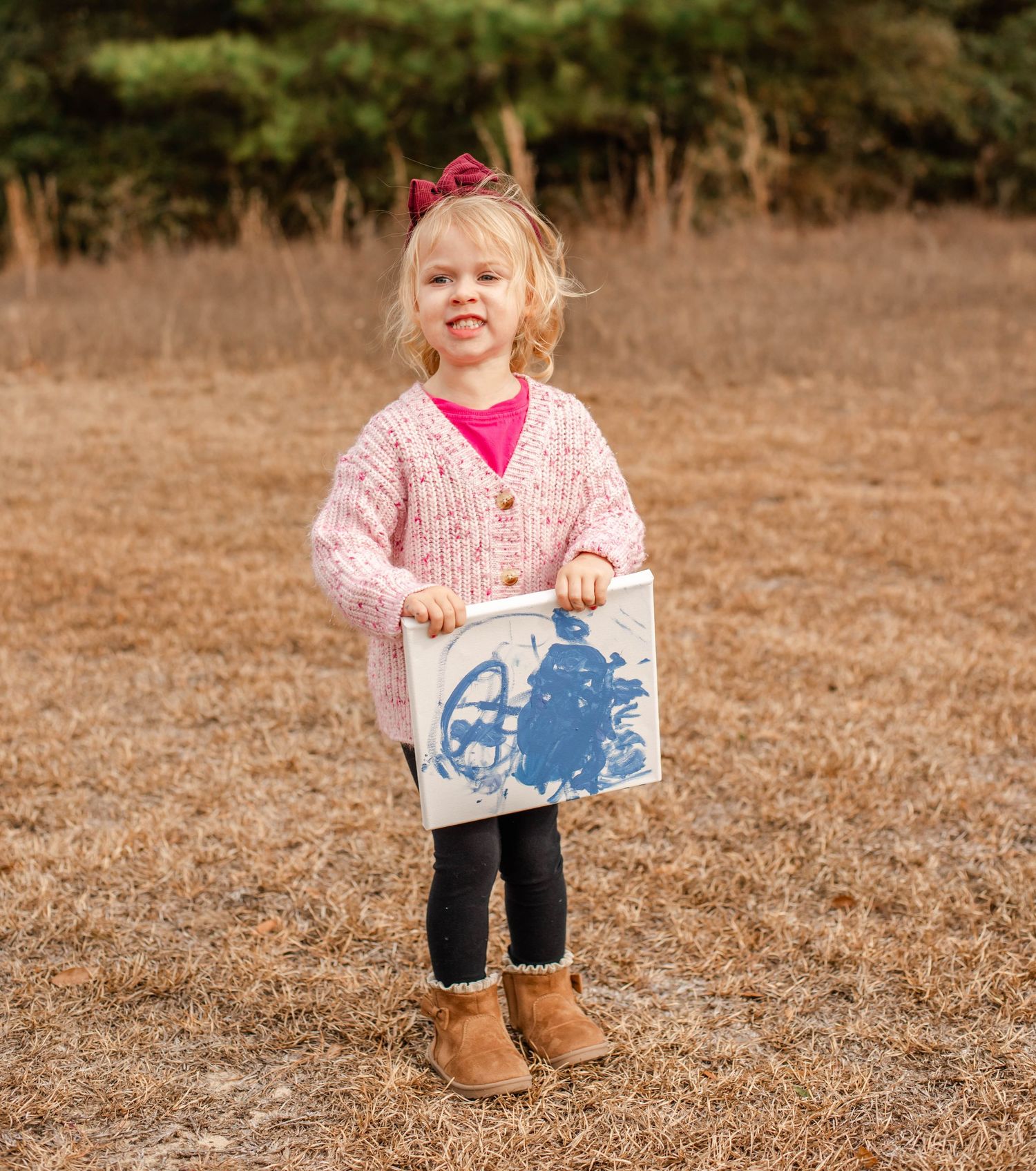 A smiling toddler girl in a pink cardigan holds a blue artwork print while standing in a dry grass field outdoors.