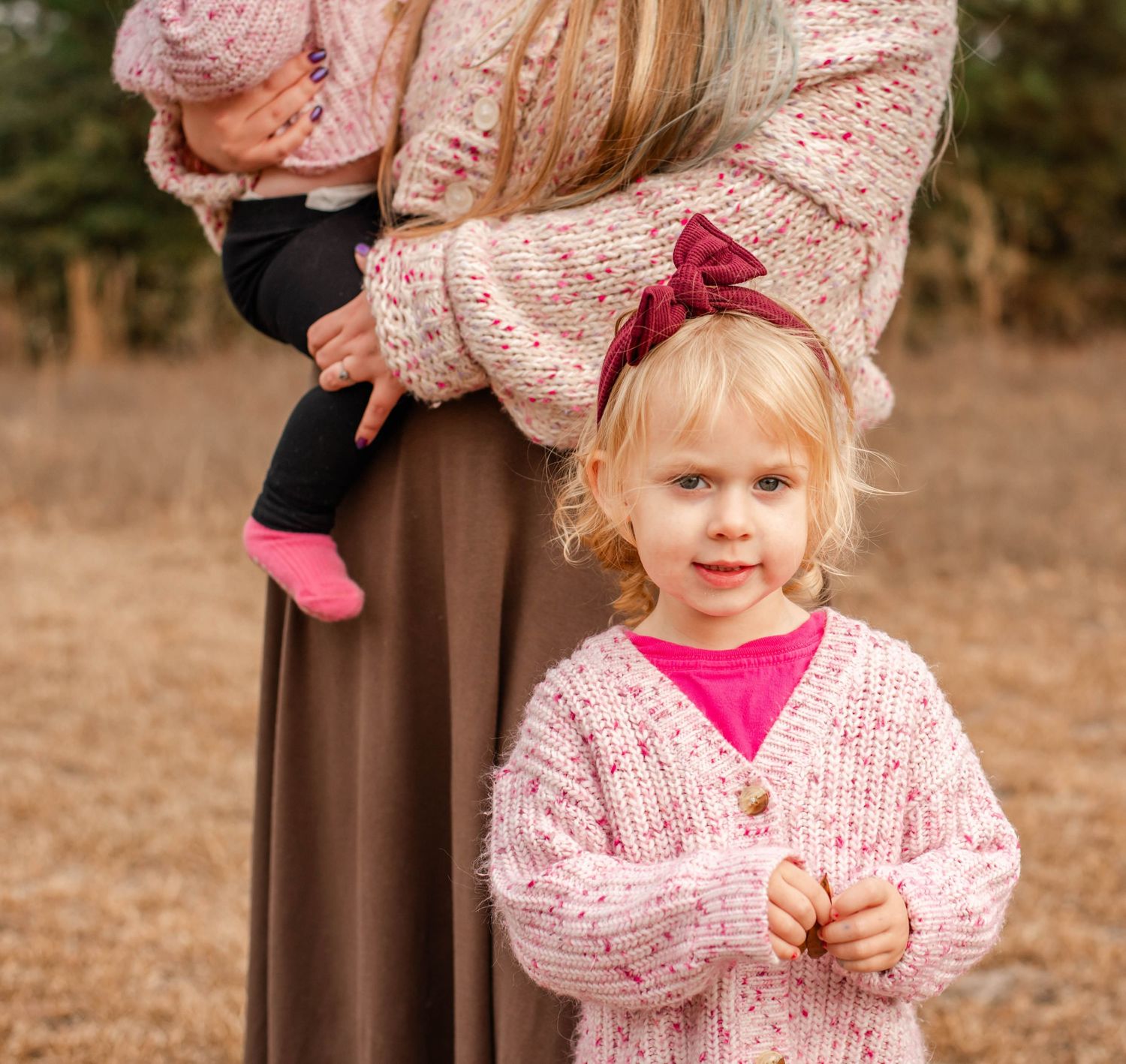 Young girl in pink knit cardigan and bow headband stands outdoors while woman holds baby behind her.